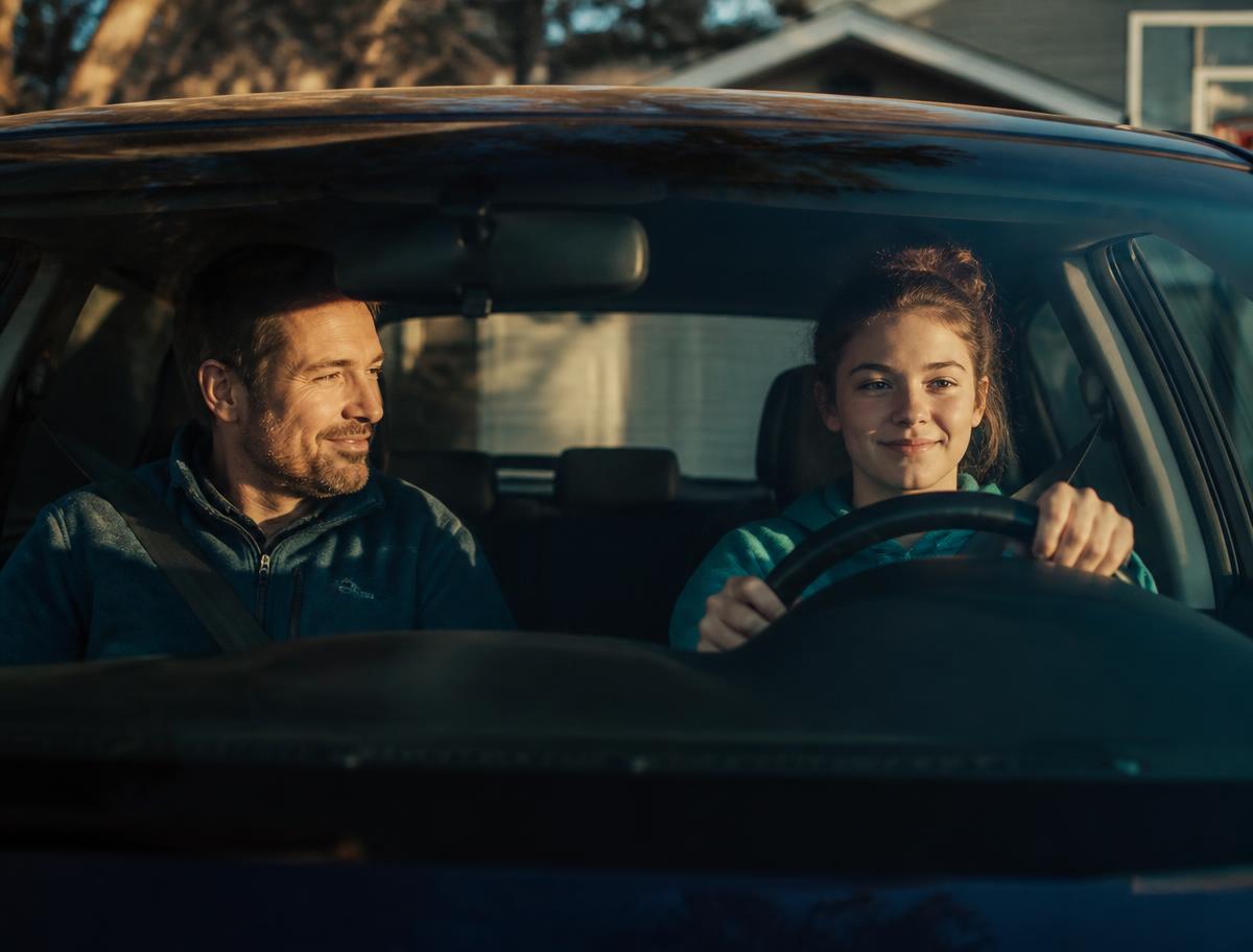 A parent riding in the passenger seat while their teenager drives in the family driveway at sunrise